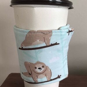 May include: A white paper coffee cup with a black lid, wrapped with a light blue fabric sleeve. The sleeve features a repeating pattern of cartoon sloths on branches. The background is a light colour.