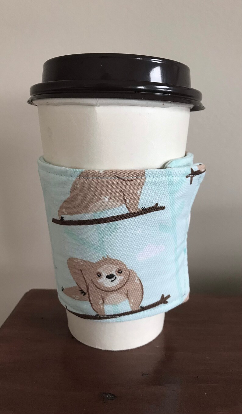 May include: A white paper coffee cup with a black lid, wrapped with a light blue fabric sleeve. The sleeve features a repeating pattern of cartoon sloths on branches. The background is a light colour.
