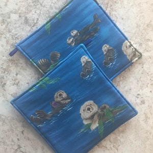 Könnte beinhalten: Zwei blaue Topflappen mit einem Muster von Seeottern, die im Meer schwimmen. Die Otter sind braun und weiß und haben süße Gesichter. Die Topflappen sind quadratisch und haben einen weißen Rand.