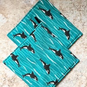 Può includere: Due presine da cucina blu turchese con un motivo di balena orca in bianco e nero. Le presine hanno anelli di sospensione neri.