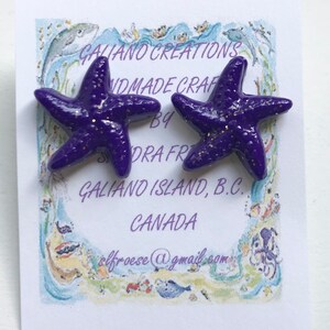 May include: A pair of purple starfish earrings with gold glitter accents. The earrings are displayed on a card with the text "Galiano Creations Handmade Crafts by Sandra Froese Galiano Island, B.C. CANADA". The email address elfroese@gmail.com is also visible.