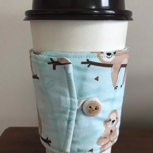 May include: A white paper coffee cup with a black lid, wrapped with a light blue fabric coffee sleeve. The sleeve features a repeating pattern of brown sloths on branches. The sleeve is secured with a button.