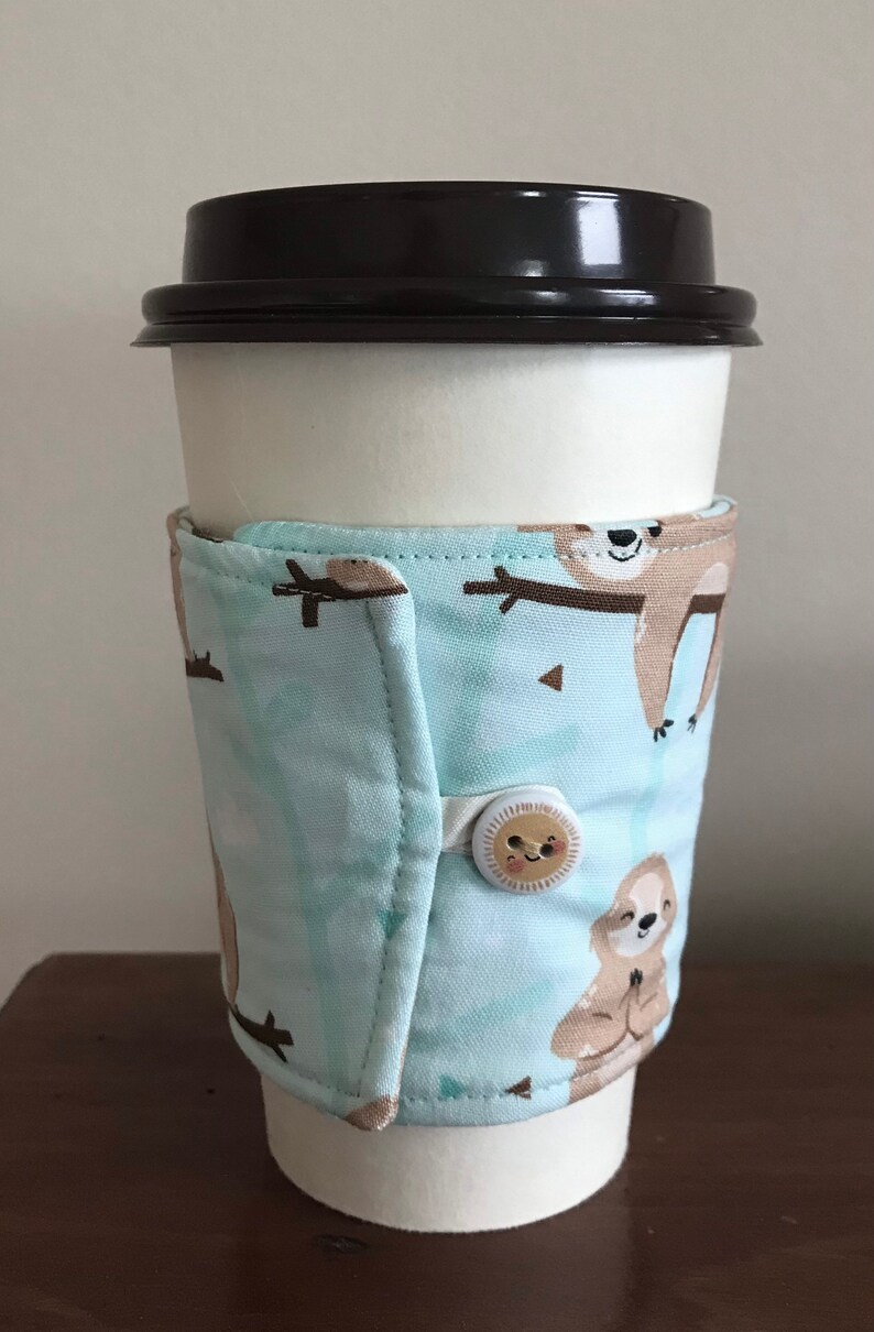 May include: A white paper coffee cup with a black lid, wrapped with a light blue fabric coffee sleeve. The sleeve features a repeating pattern of brown sloths on branches. The sleeve is secured with a button.