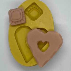 May include: A yellow silicone mold with three cavities, including a square, a diamond, and a heart shape. Two light brown soap pieces are visible: a square and a heart with a heart-shaped cutout. The mold and soap pieces are on a white surface.