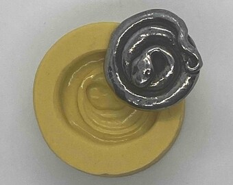 May include: A detailed image of a dark gray metal snake coiled on top of a yellow, circular mold. The snake's head is visible, and the mold has a textured, concentric design. The background is plain white.
