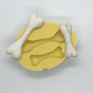 May include: A yellow silicone mold with three bone-shaped cavities. Two white bone-shaped objects are placed on top of the mold. The bones have a rounded shape with a slight curve and are off-white in color.