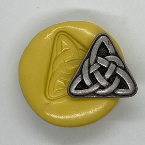 May include: A silver-colored Celtic knot charm in a triangular shape, next to a yellow wax seal with a matching embossed design. The charm has a detailed, interwoven pattern.