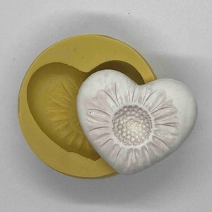 May include: A heart-shaped soap mold with a yellow exterior and a white soap heart with a floral design. The soap heart has a detailed, embossed flower pattern in a soft pink hue.