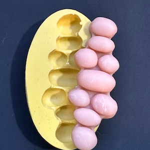 May include: A yellow silicone mold with oval-shaped cavities next to a cluster of pink, oval-shaped objects. The mold is egg-shaped, and the pink objects appear to be a cast of the mold's cavities. The background is a dark blue.