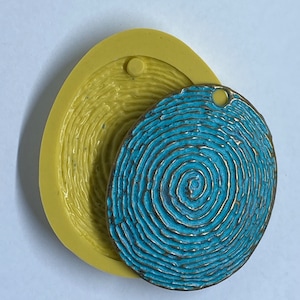 May include: A blue and gold pendant with a fingerprint design next to a yellow mold. The pendant has a hole at the top for a chain. The mold is oval-shaped and has a matching fingerprint pattern.