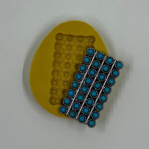 May include: A silver-toned barrette with turquoise-colored stones arranged in diagonal rows rests next to a yellow mold. The barrette has a detailed, ornate design. The mold has a pattern of small circles.