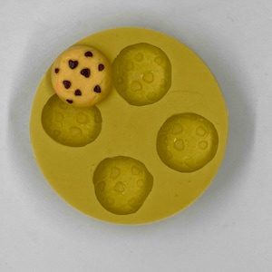 May include: A yellow silicone mold with five circular indentations, one filled with a small chocolate chip cookie. The cookie is light brown with dark brown chocolate chips. The mold is on a white surface.