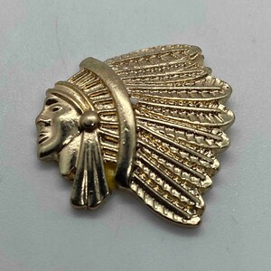 May include: Gold-toned metal pin in the shape of a Native American chief's head, featuring a detailed headdress. The pin has a smooth, polished finish and is set against a light background. The design includes facial features and feather details.