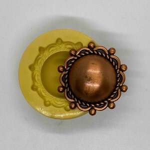 May include: A copper-colored, round button with a decorative rope-like border and small, round accents. Next to it is a yellow silicone mold with a matching design. The button has a slightly domed center.