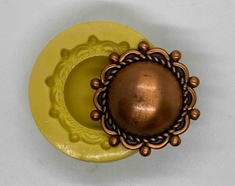 May include: A copper-colored, round button with a decorative rope-like border and small, round accents. Next to it is a yellow silicone mold with a matching design. The button has a slightly domed center.