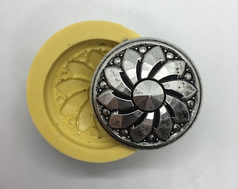 May include: A silver-colored, flower-shaped button with a faceted center and a detailed border sits next to a yellow mold. The button has a vintage, antique appearance. The mold is circular with a floral design.