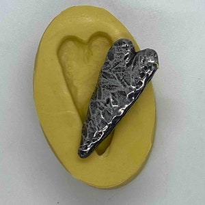 May include: A textured, silver-colored heart-shaped pendant rests on a yellow, oval-shaped mold. The pendant has a detailed, rough surface. The mold has a heart-shaped indentation. The background is a solid, light color.