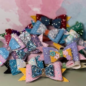 Puede incluir: Una colección de lazos para el cabello de varias capas con diversas texturas y colores brillantes, incluidos rosa, azul, morado, amarillo y verde. Varios lazos están adornados con pequeños detalles florales en el centro, mientras que otros presentan acabados de purpurina sólida y bucles de tela superpuestos.