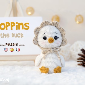 May include: A white crocheted duck doll with a beige scalloped bonnet, orange beak, and orange feet. Beside it, a white sign displays the text 'POPPins the DUCK Pattern' with American, Spanish, and French flag icons and a small pig logo.
