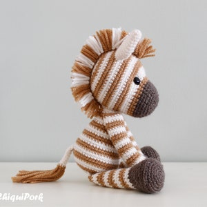 May include: A brown and white striped crocheted zebra toy with a brown mane and tail. The zebra is sitting on a white surface.