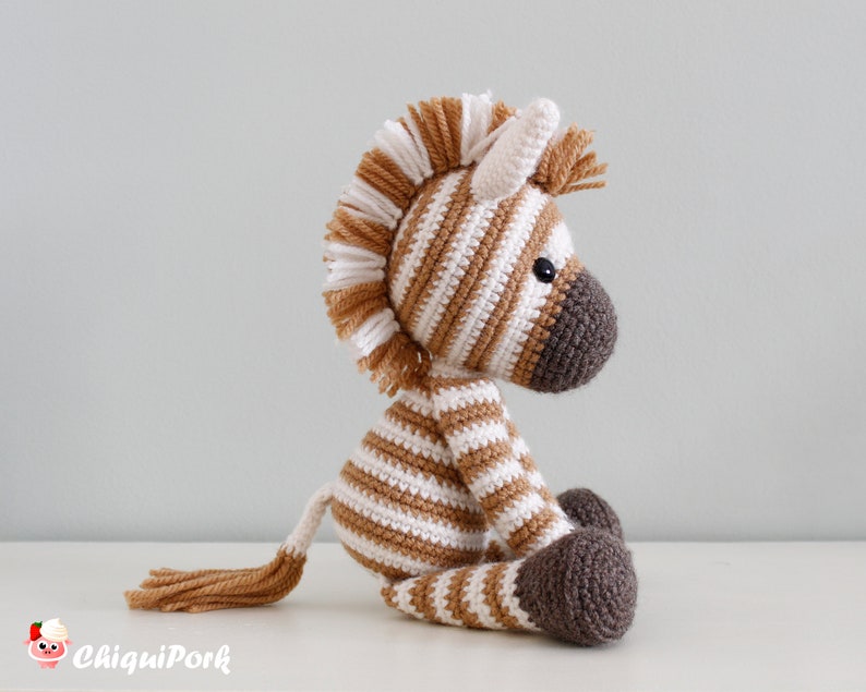 May include: A brown and white striped crocheted zebra toy with a brown mane and tail. The zebra is sitting on a white surface.