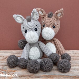 Puede incluir: Dos peluches de burro de crochet sentados juntos. El burro de la izquierda es gris con detalles blancos, y el burro de la derecha es marr&oacute;n con detalles blancos. Ambos burros tienen ojos negros y pezu&ntilde;as marrones.
