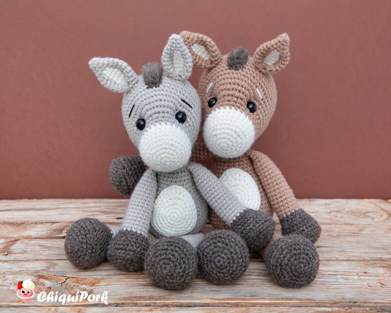 Puede incluir: Dos peluches de burro de crochet sentados juntos. El burro de la izquierda es gris con detalles blancos, y el burro de la derecha es marr&oacute;n con detalles blancos. Ambos burros tienen ojos negros y pezu&ntilde;as marrones.