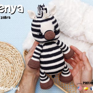 May include: Kenya the zebra is a crocheted stuffed animal toy. The zebra is black and white with brown hooves and a black nose. The zebra is being held by a person's hands.
