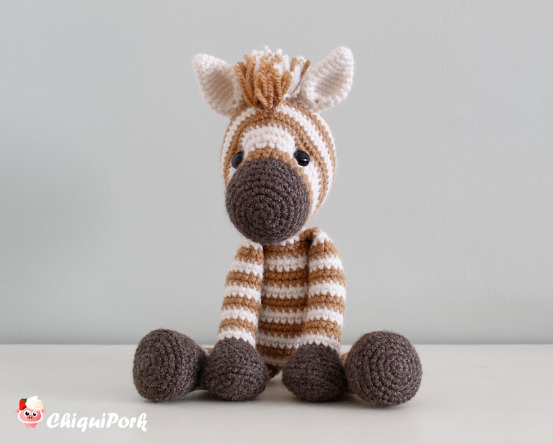 May include: A crocheted zebra stuffed animal with brown and white stripes. The zebra is sitting on a white surface.