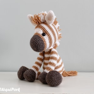 May include: A crocheted zebra stuffed animal with brown and white stripes. The zebra has a brown nose and black eyes. The zebra is sitting on a white surface.