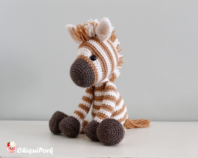 May include: A crocheted zebra stuffed animal with brown and white stripes. The zebra has a brown nose and black eyes. The zebra is sitting on a white surface.