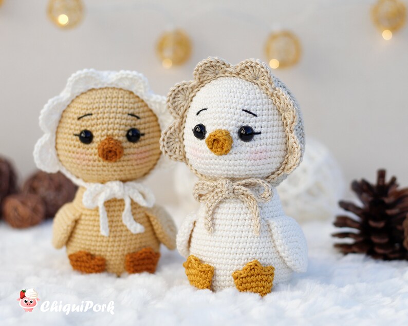 May include: Two crocheted ducklings with black eyes and pink cheeks. The left duckling is light brown with a white bonnet, and the right duckling is white with a beige bonnet. Both have orange feet and beaks. The text ChiquiPork is in the corner.