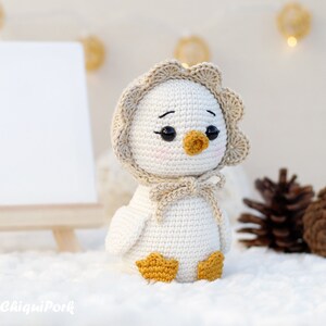 May include: A small, off-white crocheted duckling figurine featuring a scalloped-edge bonnet in a light beige hue, tied with a bow under the chin. The duckling has black bead eyes, a small mustard-yellow beak, and matching mustard-yellow webbed feet.