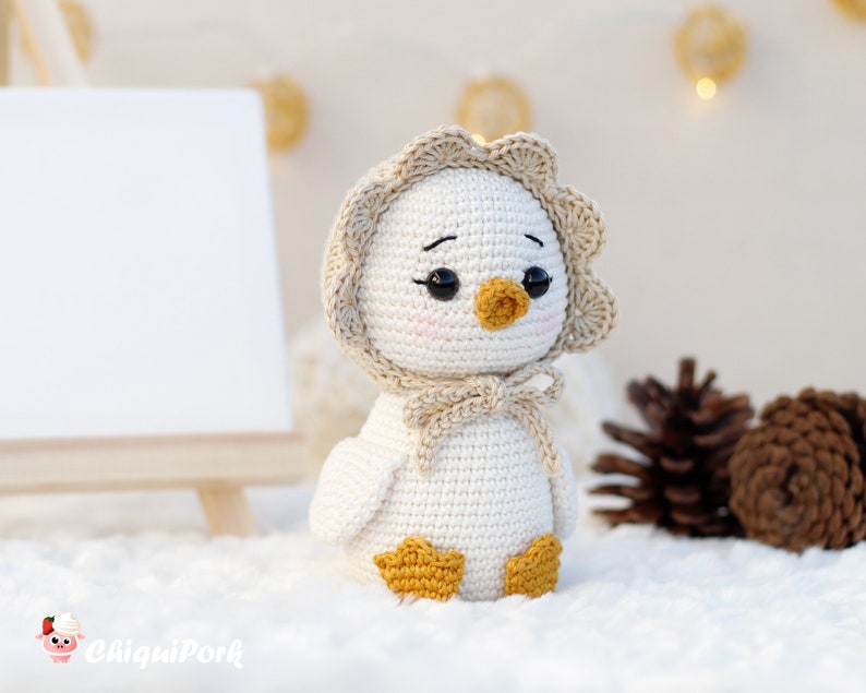 May include: A small, off-white crocheted duckling figurine featuring a scalloped-edge bonnet in a light beige hue, tied with a bow under the chin. The duckling has black bead eyes, a small mustard-yellow beak, and matching mustard-yellow webbed feet.