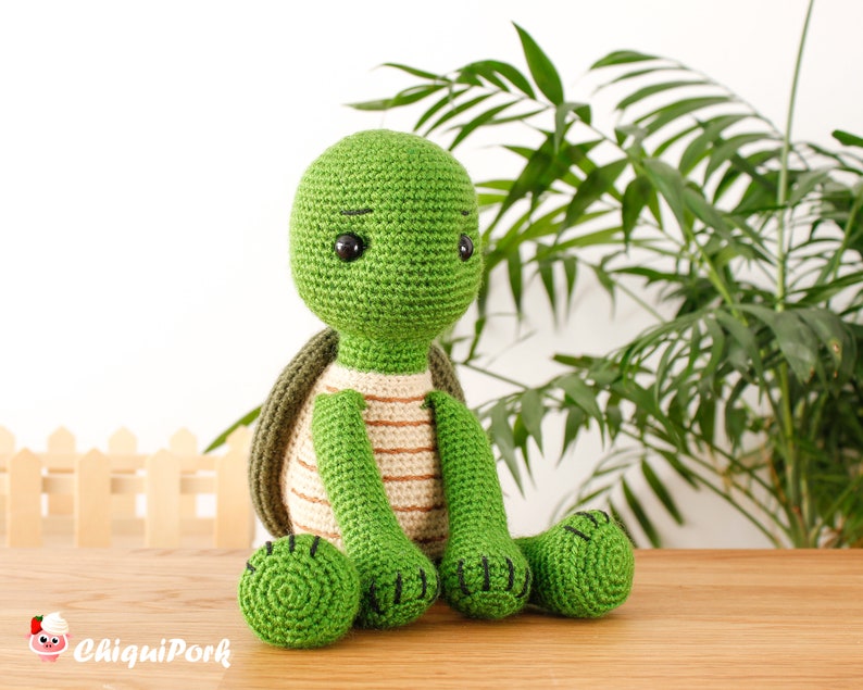 以下が含まれることがあります： 緑色の毛糸で編まれた、茶色の縞模様の甲羅を持つ、ぬいぐるみの亀。亀は木の表面に座っており、背景には緑色の植物がある。