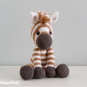 May include: A crocheted zebra stuffed animal with brown and white stripes. The zebra has a brown nose and black eyes. The zebra is sitting on a white surface.