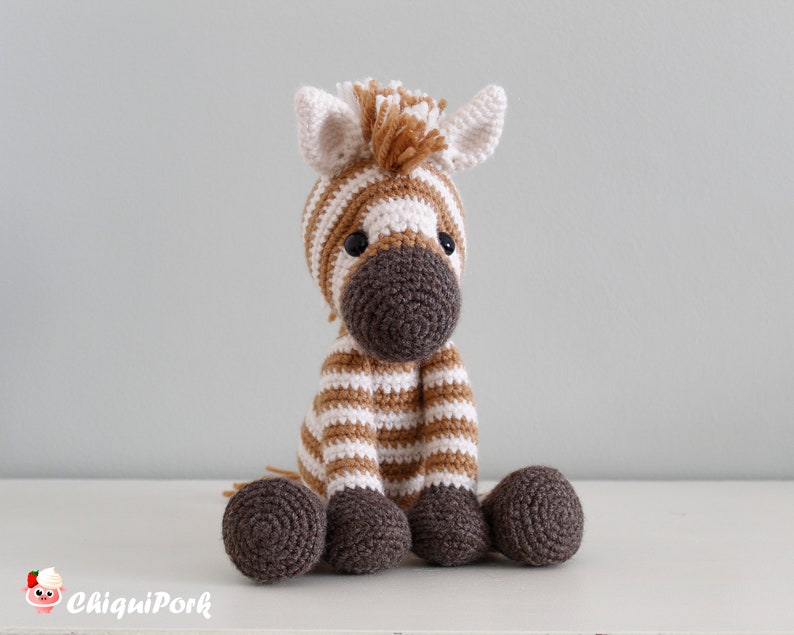May include: A crocheted zebra stuffed animal with brown and white stripes. The zebra has a brown nose and black eyes. The zebra is sitting on a white surface.