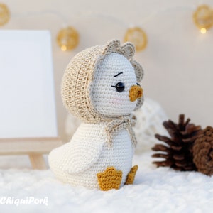 May include: A white crocheted duckling toy featuring a beige bonnet with a scalloped edge tied under the chin, black safety eyes, a small orange beak, and orange webbed feet. The toy has a smooth, rounded body and a soft, textured appearance.
