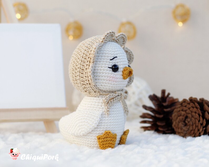May include: A white crocheted duckling toy featuring a beige bonnet with a scalloped edge tied under the chin, black safety eyes, a small orange beak, and orange webbed feet. The toy has a smooth, rounded body and a soft, textured appearance.