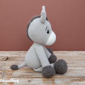 Puede incluir: Un burro de crochet gris sentado en una superficie de madera. El burro tiene una melena y una cola marrones, y ojos negros. El burro est&aacute; mirando hacia la derecha.