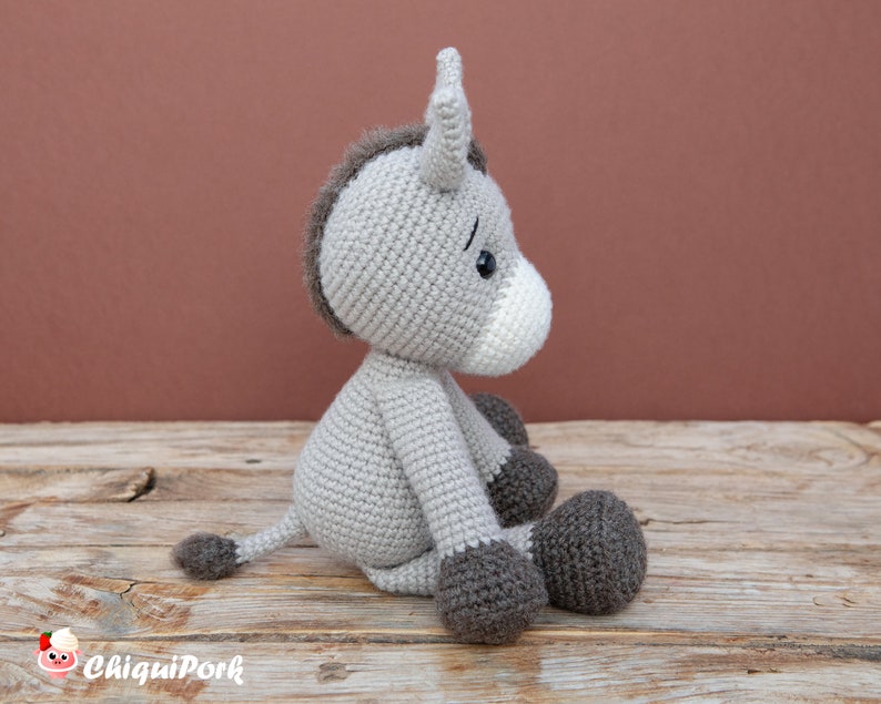 Puede incluir: Un burro de crochet gris sentado en una superficie de madera. El burro tiene una melena y una cola marrones, y ojos negros. El burro est&aacute; mirando hacia la derecha.