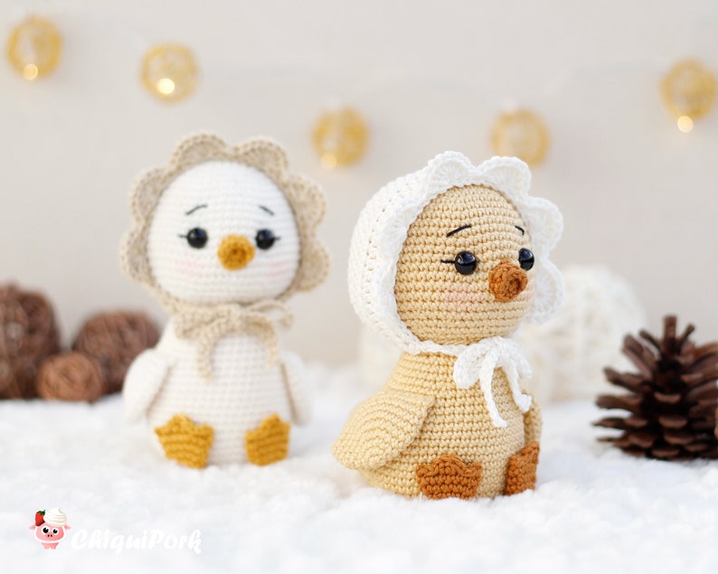 May include: Two crocheted ducklings with black bead eyes and pink cheeks. The left duckling is white with a beige bonnet, and the right duckling is light brown with a white bonnet. Both have orange feet and beaks, and the text "ChiquiPork" is visible.