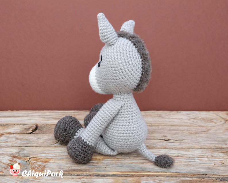Puede incluir: Un juguete de peluche de burro de crochet gris sentado en una superficie de madera. El burro tiene detalles marrones en sus pezu&ntilde;as, cola y crin.