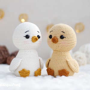 May include: Two crocheted ducklings, one white and one light tan, featuring black plastic eyes, embroidered eyebrows, and pink blush on their cheeks. The white duck has a yellow beak and feet, while the tan duck has a brown beak and feet.