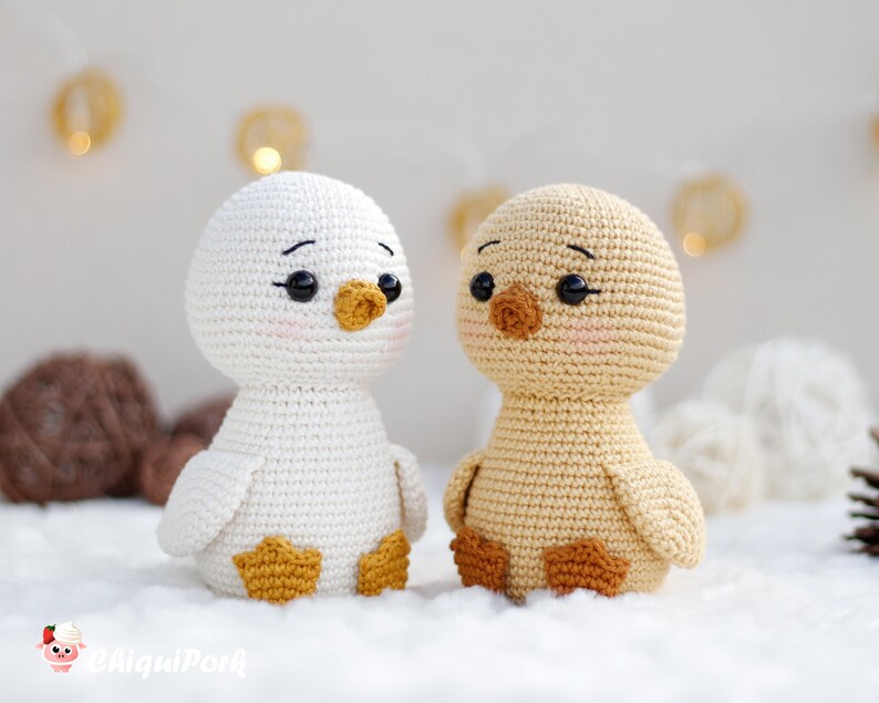 May include: Two crocheted ducklings, one white and one light tan, featuring black plastic eyes, embroidered eyebrows, and pink blush on their cheeks. The white duck has a yellow beak and feet, while the tan duck has a brown beak and feet.