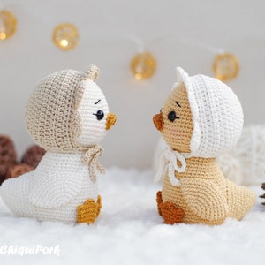 May include: Two crocheted ducklings facing each other. The left duckling is white with a beige bonnet and orange feet. The right duckling is beige with a white bonnet and orange feet. Both have black eyes, pink cheeks, and the text ChiquiPork.