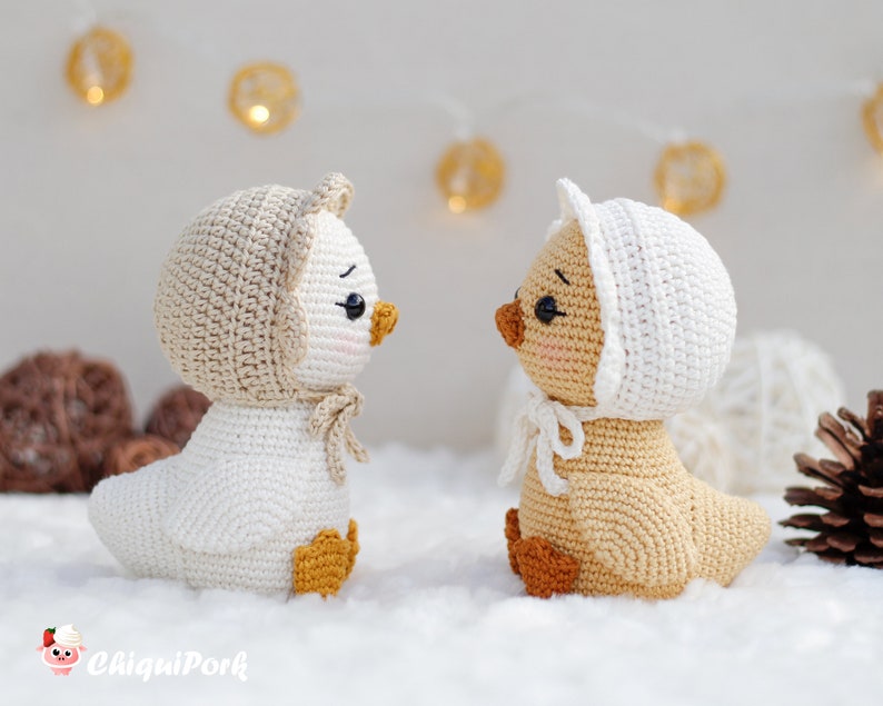 May include: Two crocheted ducklings facing each other. The left duckling is white with a beige bonnet and orange feet. The right duckling is beige with a white bonnet and orange feet. Both have black eyes, pink cheeks, and the text ChiquiPork.