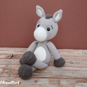 Puede incluir: Un juguete de burro de crochet gris con pezu&ntilde;as marrones, detalles blancos y ojos negros. El burro est&aacute; sentado sobre una superficie de madera.