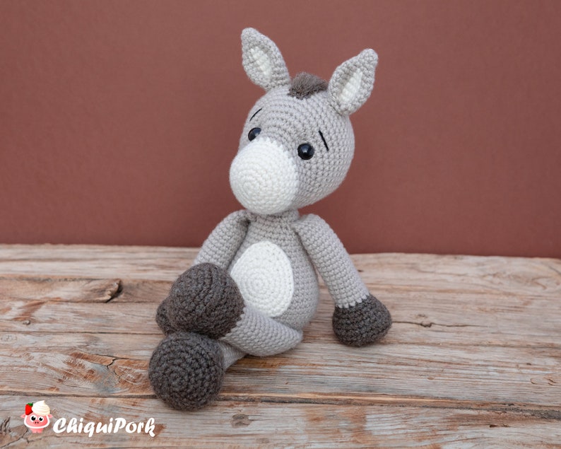 Puede incluir: Un juguete de burro de crochet gris con pezu&ntilde;as marrones, detalles blancos y ojos negros. El burro est&aacute; sentado sobre una superficie de madera.