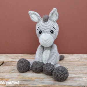 Puede incluir: Un juguete de peluche de burro de crochet gris con ojos negros y nariz blanca. El burro est&aacute; sentado sobre una superficie de madera.